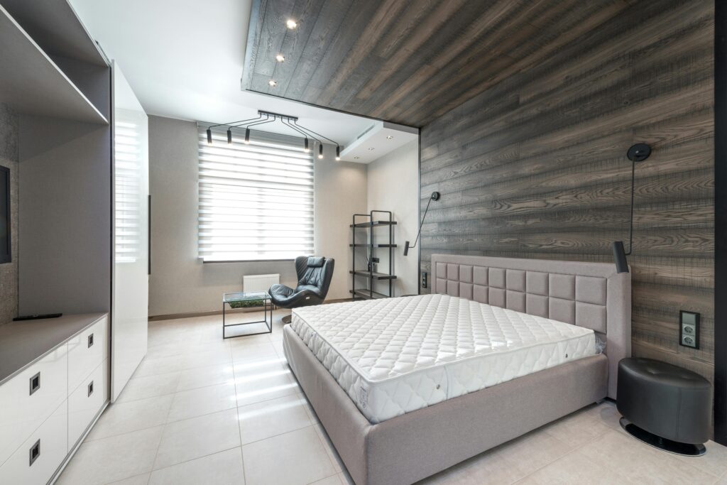 Bed with white mattress placed at wall with decorative element against cupboards in light stylish bedroom with window and armchair